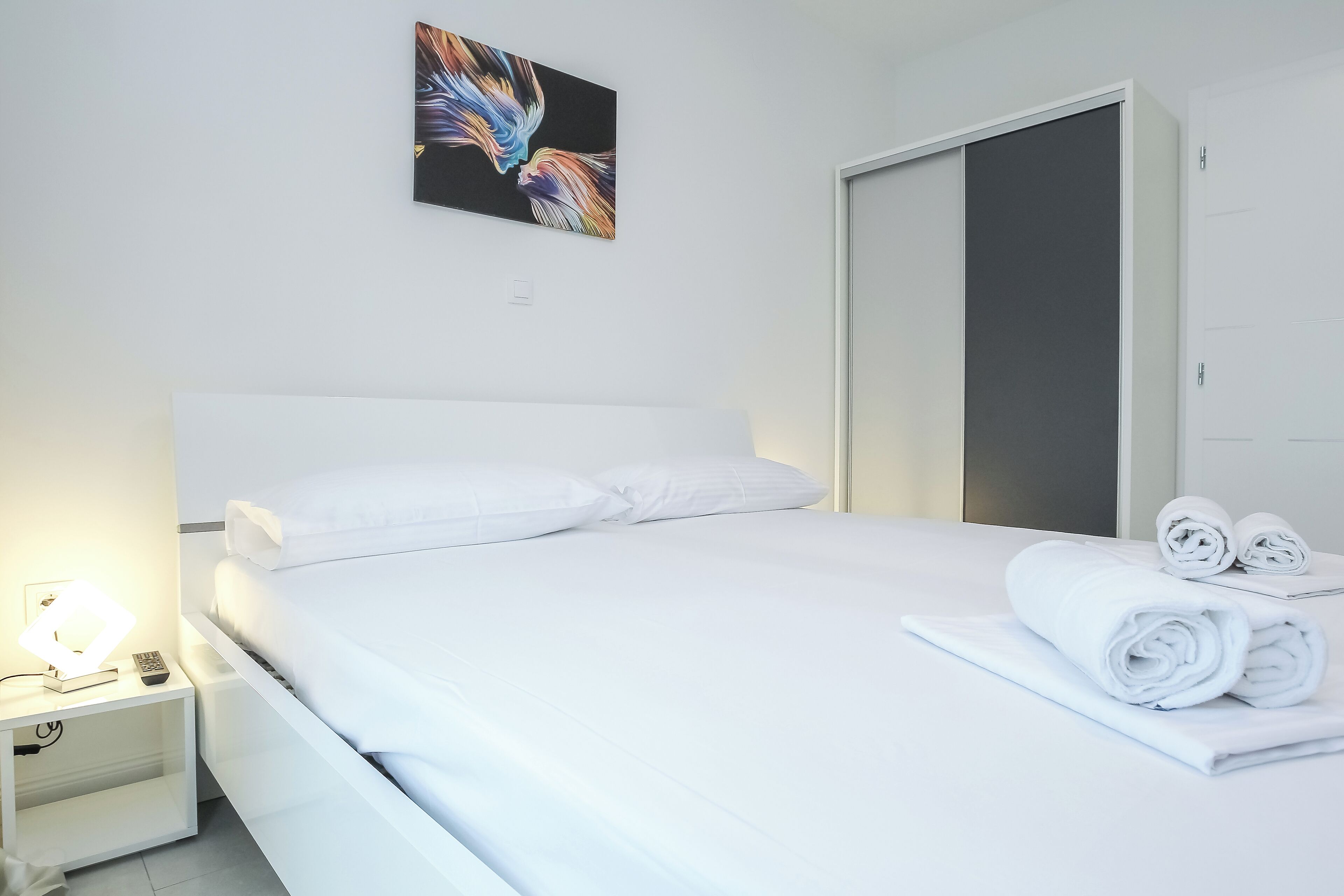 A neatly made bed with white linens and pillows, a bedside lamp, a folded towel, and a painting on the wall.