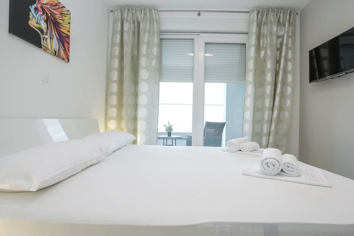 Habitación de hotel con una cama grande, cortinas blancas, un televisor montado en la pared y vistas al balcón exterior.