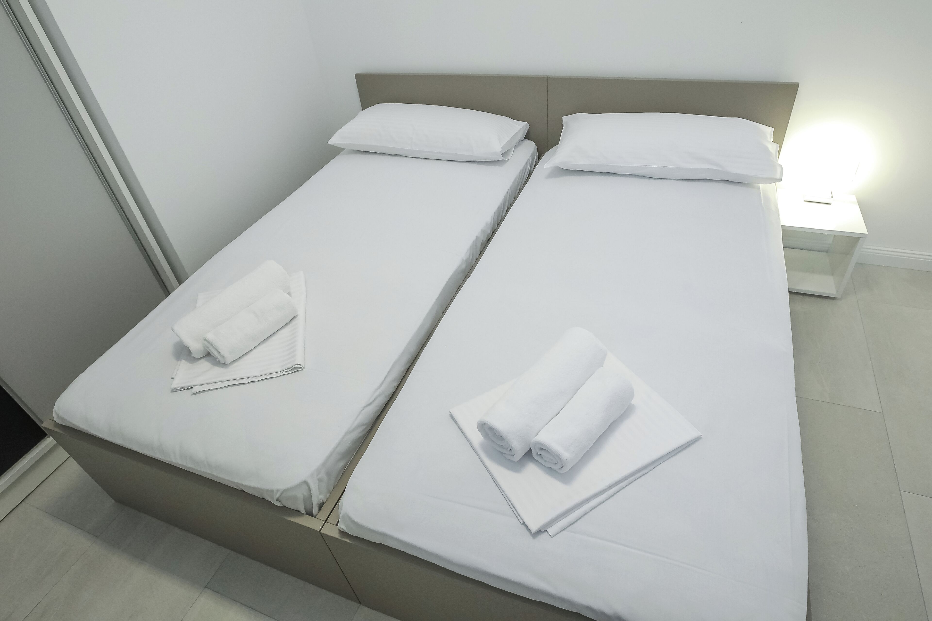 Two single beds with white linens and towels, a bedside table with a lamp, and a plain wall.