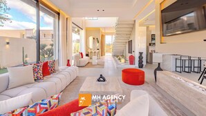 A modern living room with a sofa, coffee table, and red ottoman. There is a TV mounted on the wall and a staircase leading to another level. - Villa Akenza (Marrakech)