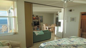A room with a bed, a sofa, a dining table with chairs, a bookshelf, and a view of the sea through a window. - Estero Beach & Tennis Club 1105A! Gorgeous Direct Gulf View! (Fort Myers Beach)