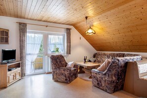A cozy room with a wooden ceiling, a television, a sofa, a table with bottles, and a window with curtains.