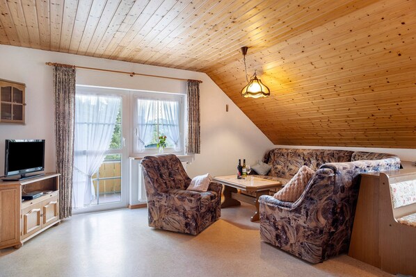 Une pièce chaleureuse avec un plafond en bois, une télévision, un canapé, une table avec des bouteilles et une fenêtre avec des rideaux.