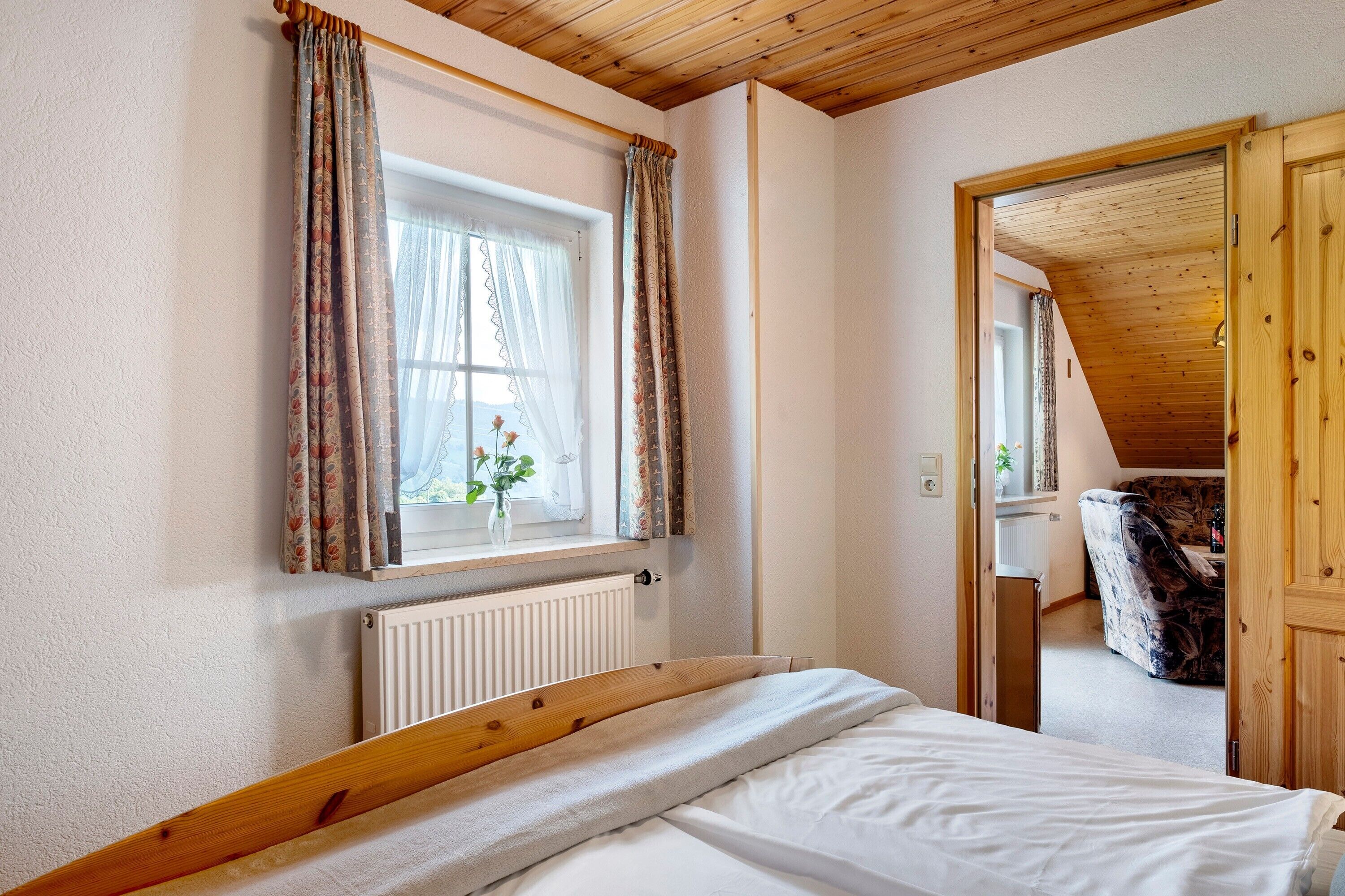 A bedroom with a wooden headboard, a window with curtains, a radiator, and a view into another room.