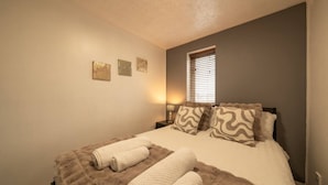 Un dormitorio con una cama, dos mesitas de noche, un ventanal con persianas y tres cuadros en la pared.