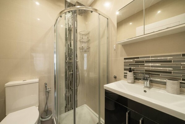 Un baño moderno con ducha envuelta en vidrio, un lavabo blanco y un dispensador de jabón negro, y un salpicadero de azulejos.