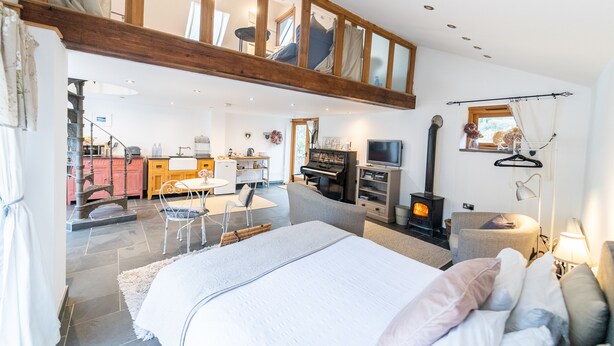 Ett rymligt sovrum med en stor säng, en träinredd loftvåning ovanför, en öppen spis och en matplats med ett piano.