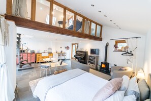 A spacious bedroom with a large bed, a wooden loft above, a fireplace, and a dining area with a piano. - Stunning riverside & waterfall apartment barn, nestled in nature Brecon Beacons (Llangynidr)
