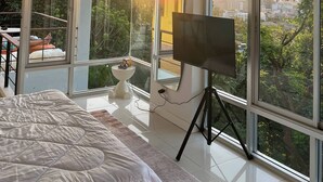 A bedroom with a bed, a TV on a tripod, a small table, and large windows with a city view. - Jungle House Phuket - Seaview Retreat (Patong)