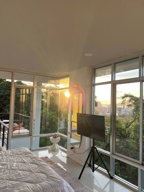 Une chambre à coucher avec un lit, un téléviseur posé sur un trépied, une petite table et de grandes fenêtres donnant sur la ville.