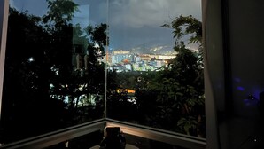 A room with a large window, a round table, and a view of a cityscape at dusk. - Jungle House Phuket - Seaview Retreat (Patong)