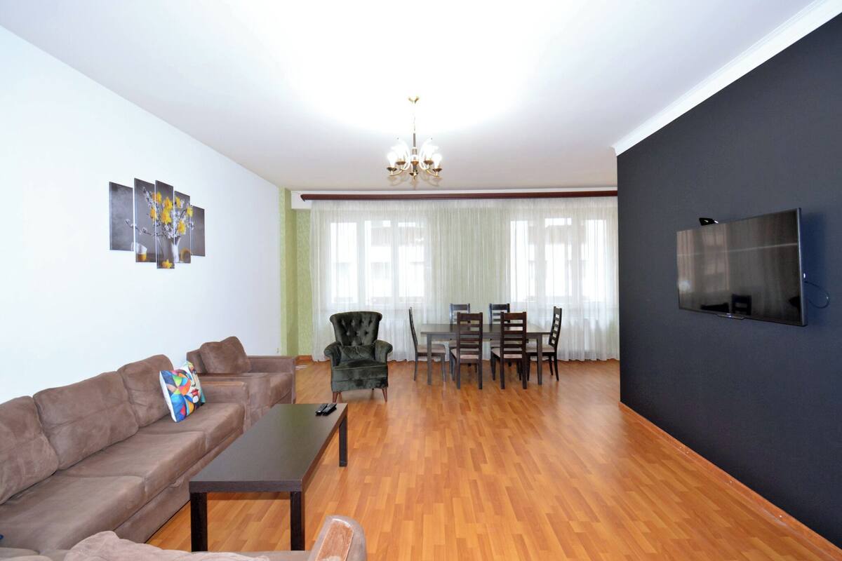 Ein Wohnzimmer mit einer Couch, einem Esstisch mit Stühlen, einem an der Wand befestigten Fernseher und einer Deckenleuchte.