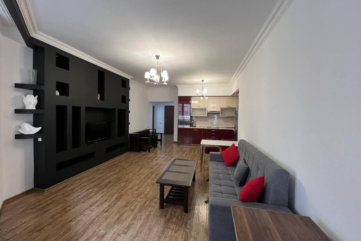 Ein modernes Wohnzimmer mit dunkler Akzentwand, einem grauen Sofa mit roten Kissen, einem Holztisch und einem Essbereich mit dunklem Tisch und Stühlen.