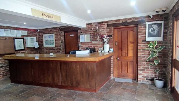 A reception area with a wooden reception desk, a sign labeled "Reception," and a brick wall with framed pictures and certificates. - Rivenhall End Hotel (Witham)