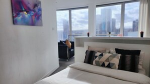 A modern hotel room with a large bed, a view of the city through large windows, and a colorful abstract painting on the wall. - Cityscape Apartment in Rotunda Bham (Birmingham)