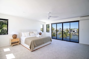 A modern bedroom with a large bed, a nightstand, a ceiling fan, and a view of a body of water through floor-to-ceiling windows.