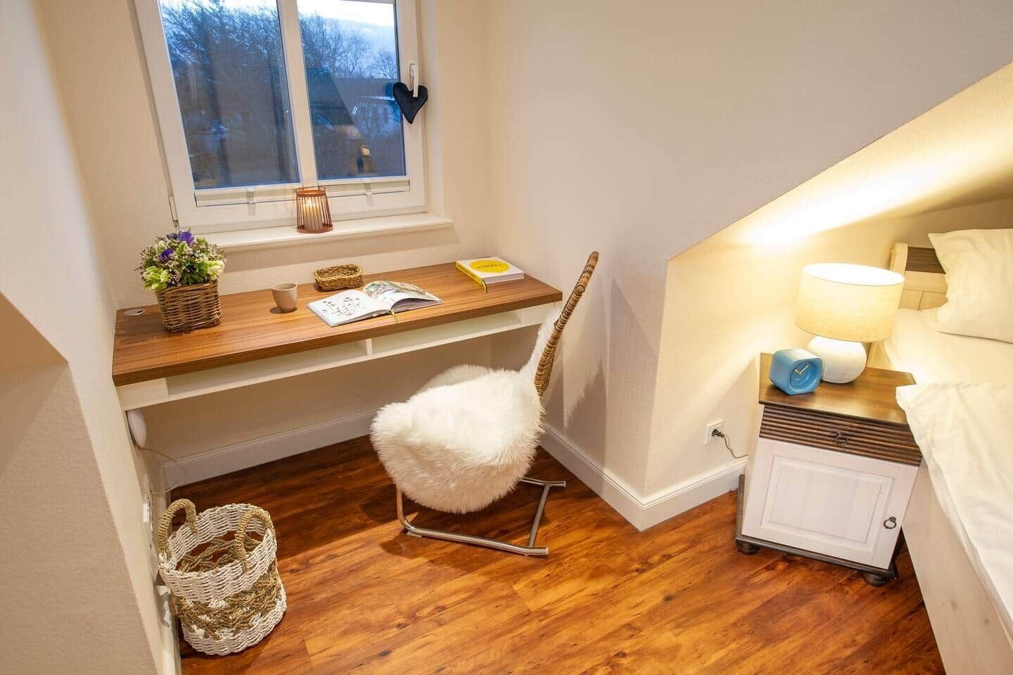 Un dormitorio acogedor con un escritorio de madera, una butaca mullida, una mesita de noche con lámpara y una ventana decorada con un adorno en forma de corazón.