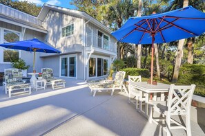 A covered outdoor patio with white furniture and blue umbrellas, surrounded by a house with white siding and a balcony.