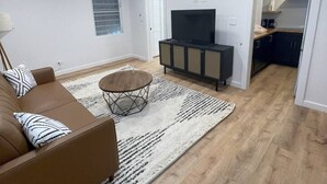 Een moderne woonkamer met een bruine bank, een ronde salontafel en een televisie op een zwarte kast. De kamer heeft houten vloeren en witte muren.
