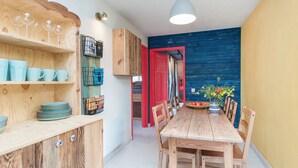 Een houten eethoek met een tafel, stoelen, een plank met drinkglazen en een rode deur naar een andere kamer.
