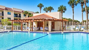 A swimming pool with lounge chairs and a covered seating area, surrounded by palm trees and hotel buildings.
