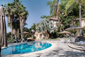 A swimming pool with lounge chairs and umbrellas, surrounded by palm trees and a building with arched windows.