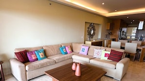 A modern living room with a beige sectional sofa, colorful pillows, a wooden coffee table, and a kitchen in the background. - Anana El Dorado 305 Condo With Pool Lake Views (Chapala)