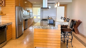 A modern kitchen with wooden cabinetry, stainless steel appliances, granite countertops, and a bar area with stools. - Anana El Dorado 305 Condo With Pool Lake Views (Chapala)