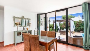 Uma área de refeições com mesa de vidro e cadeiras de vime, um armário azul claro com objetos decorativos, e vista para um pátio ao ar livre com móveis e vegetação.