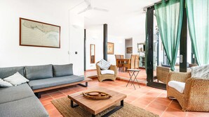 Uma sala de estar moderna com um sofá modular cinza, uma mesa de centro de madeira, cadeiras de vime e um espaço de jantar com uma mesa pequena e cadeiras.