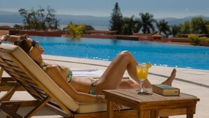 A person relaxing by a pool with a drink and a book, surrounded by palm trees and a clear view of the mountains. - Anana El Dorado 203 Condo With Lake Views Pool (Chapala)