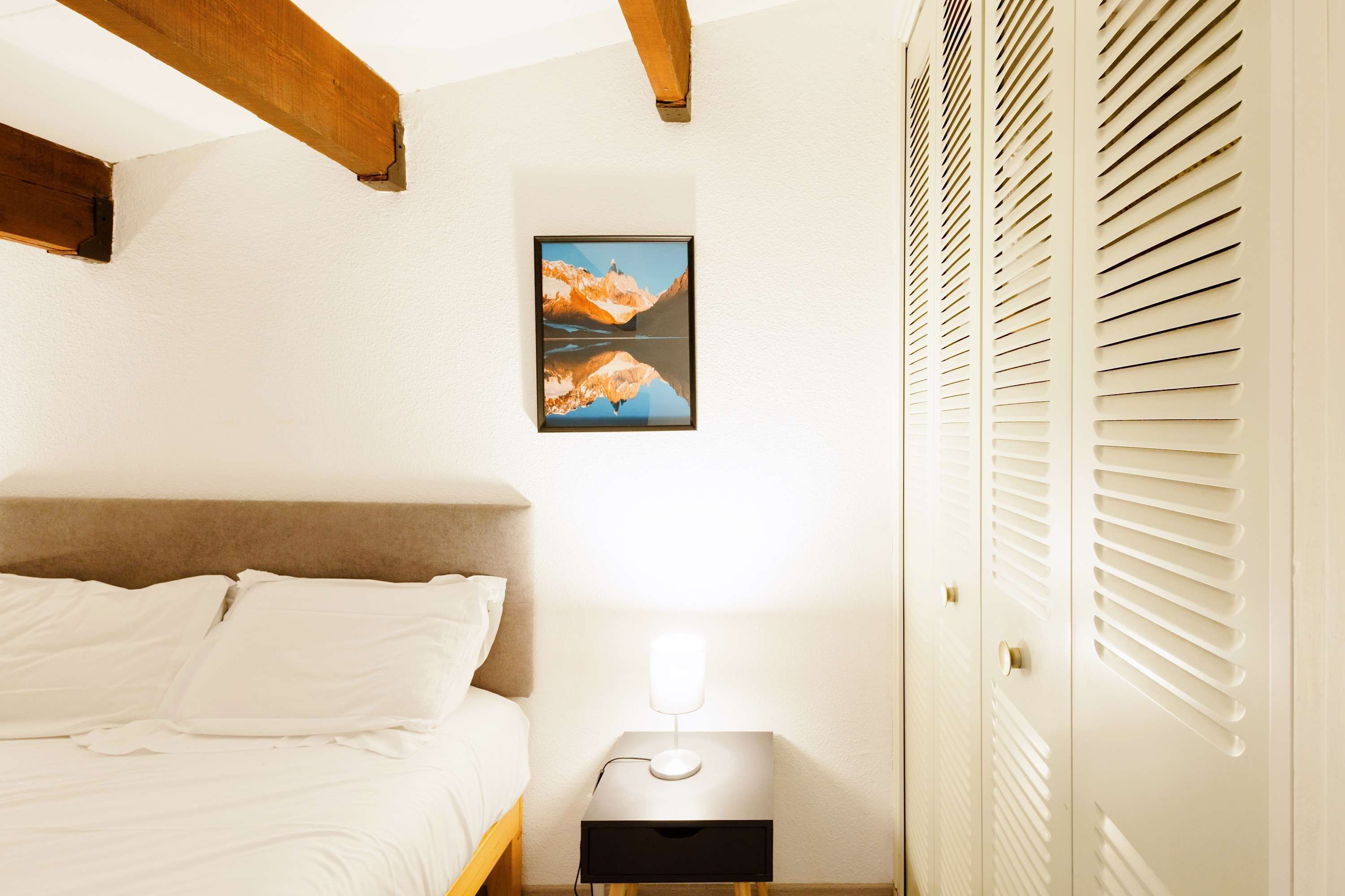 Ein Schlafzimmer mit einem Bett, einem Nachttisch mit Lampe, einem gerahmten Bild an der Wand und einem Schrank mit Klappläden.
