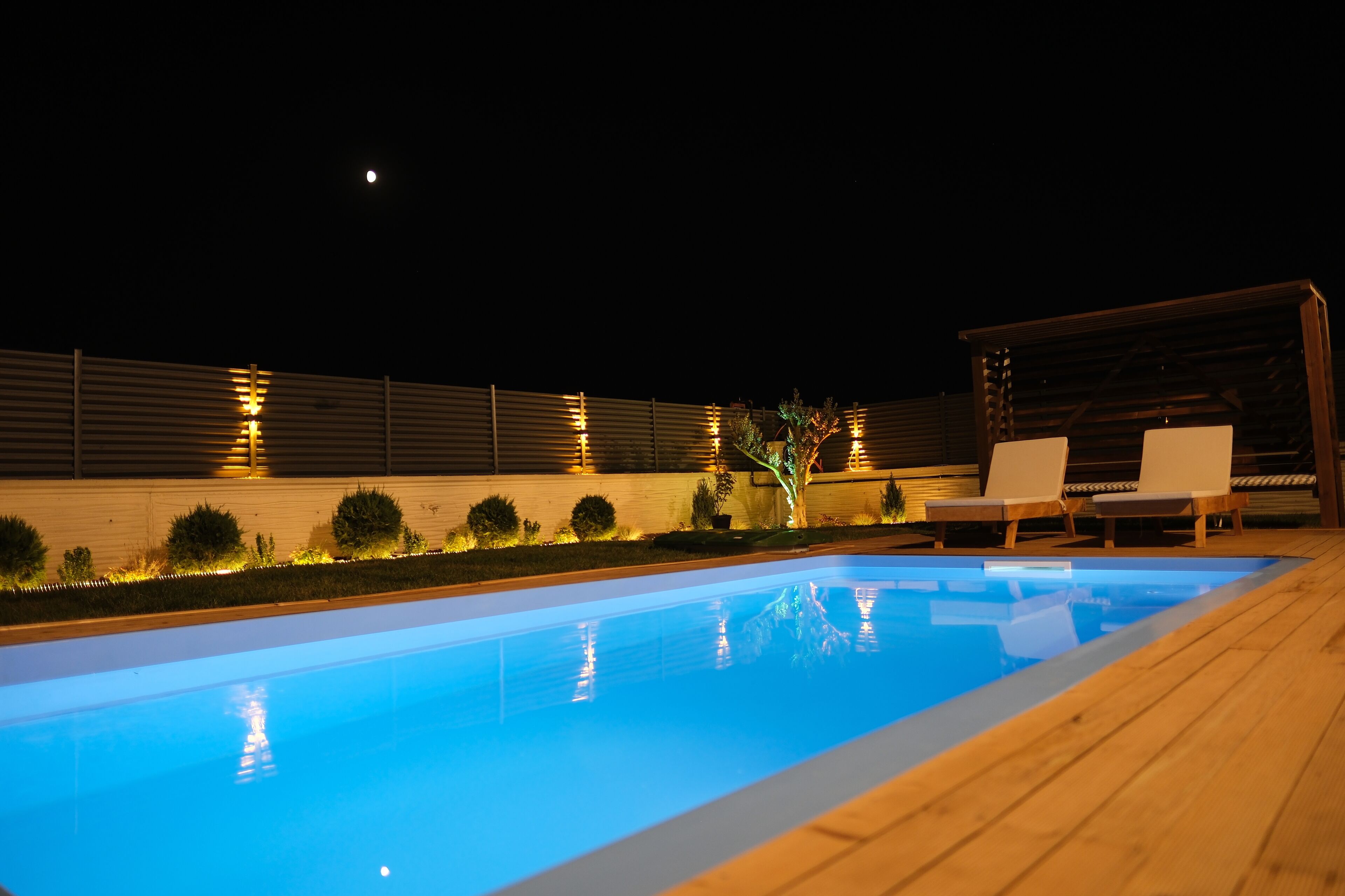 Piscina con cubierta de madera y sillas de descanso, rodeada por un muro bajo con luces.