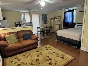 A cozy room with a brown leather sofa, a bed with a white comforter, a dining table with chairs, and a ceiling fan. - Charming studio in beautiful St. Petersburg with AC (St. Petersburg)