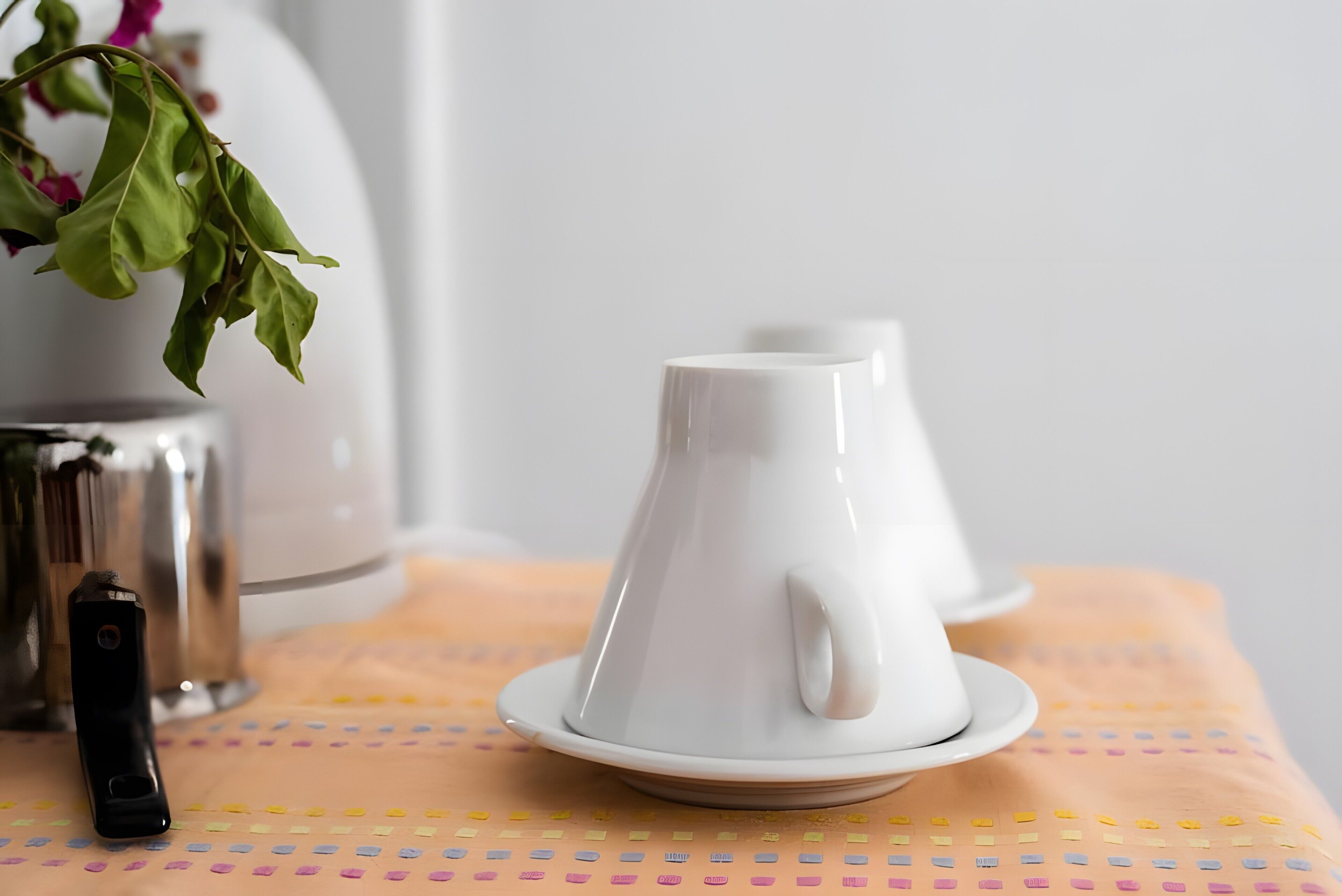 Une théière blanche posée sur une soucoupe, un objet noir sur la table et un vase avec des feuilles vertes.
