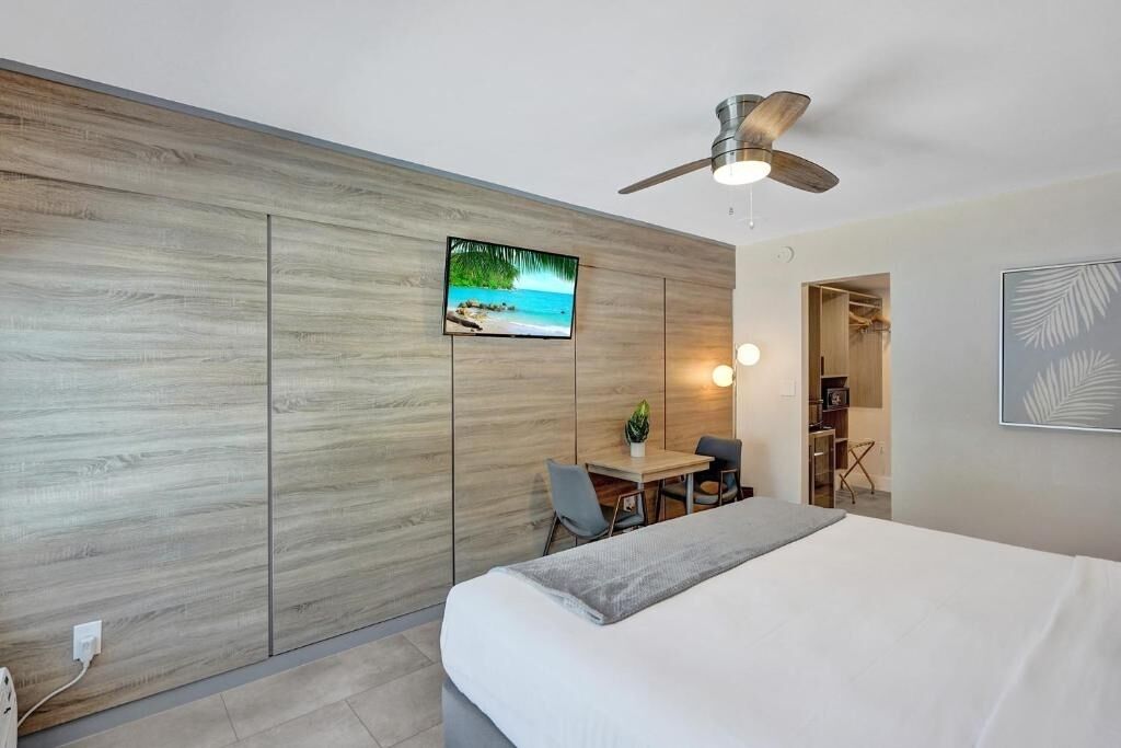 A modern hotel room with a bed, a desk, a chair, a ceiling fan, and a TV on the wall.