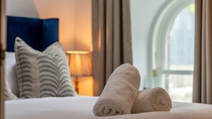 A neatly made bed with rolled towels, a lamp, and a view of a building through an arched window.