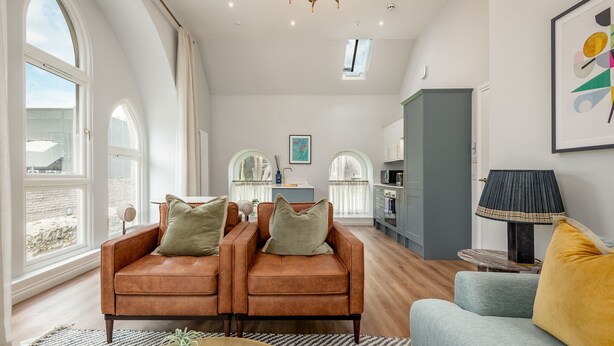 A spacious living area with a brown leather sofa set, a light blue armchair, and a round coffee table. There are large windows with curtains, a chandelier, and a framed abstract painting on the wall.