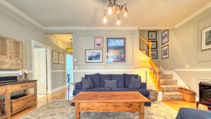 A living room with a dark blue sofa, a wooden coffee table, and a patterned rug. There are framed pictures on the wall, a staircase, and a wooden cabinet with a television.