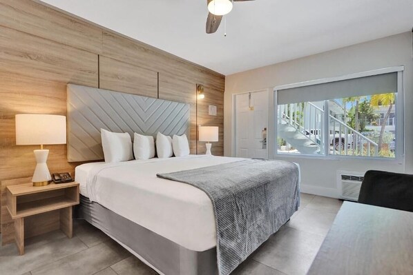 A modern bedroom with a large bed, wooden headboard, two bedside tables with lamps, and a large window with a view of the outside.