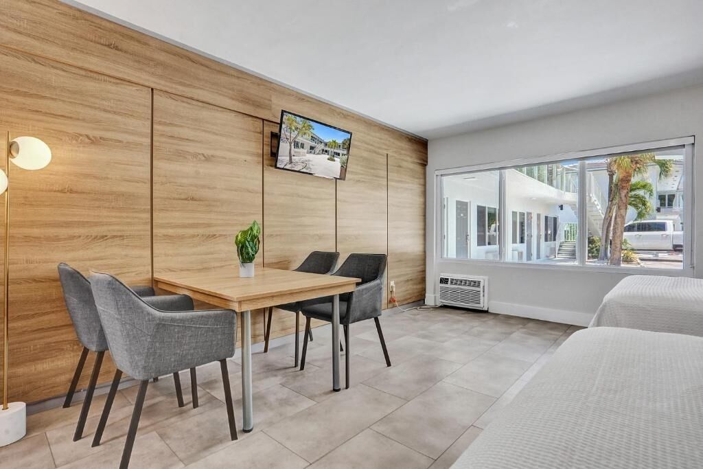 Une chambre d’hôtel moderne avec un mur d’accent en bois, un téléviseur à écran plat, une table à manger avec des chaises, une plante en pot et une grande fenêtre donnant sur l’extérieur.