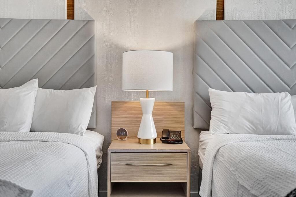 A hotel room with two beds, a nightstand with a lamp, a clock, and a phone.