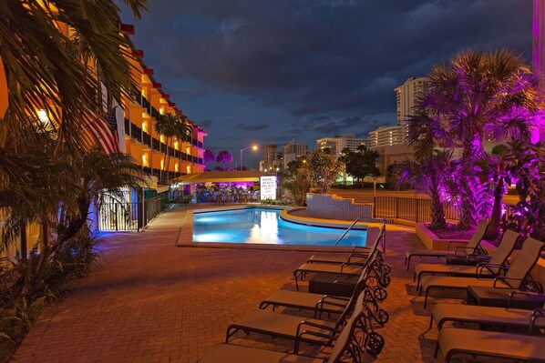 Zona de piscina de hotel con sillones de descanso, palmeras y un letrero que indica el Hotel Royal Beach.