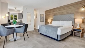 Una habitación de hotel moderna con una cama grande, un área de comedor con mesa y sillas, y una kitchenette al fondo.