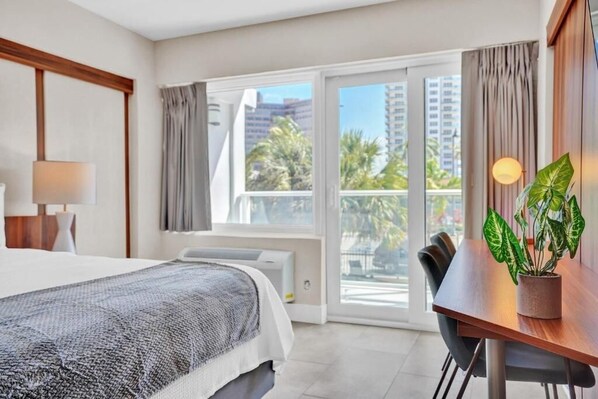 Habitación de hotel con cama, escritorio y vistas a palmeras y edificios.