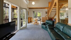 A spacious living area with a large flat-screen TV, a green leather sofa, a wooden dining table with chairs, and a staircase leading to an upper level.