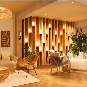 Een moderne woonkamer met een wand van houten latten, twee rieten stoelen, een witte bank en een grote potplant.