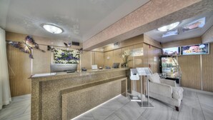 A hotel reception area with a granite counter, a floral wall decoration, a sofa, and a television screen displaying images. - Relaxing Retreat with Onsite Pool | 2 Suites Near Downtown Attractions (Fort Lauderdale)