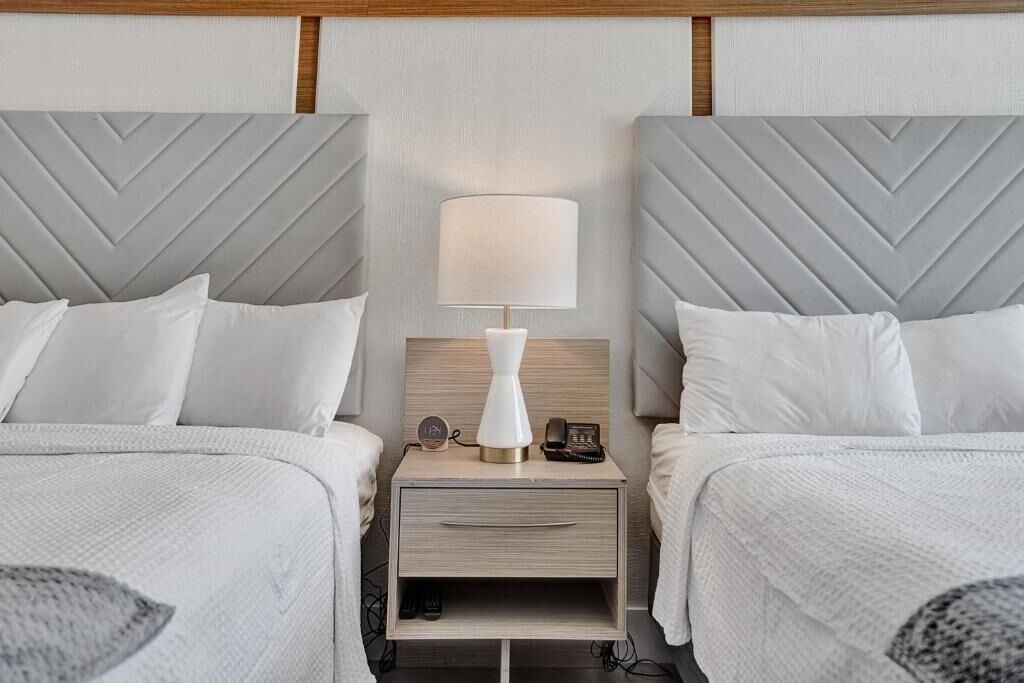 Une chambre d’hôtel avec deux lits, chacun doté d’une tête de lit grise, d’une table de chevet avec une lampe, une horloge et un téléphone.