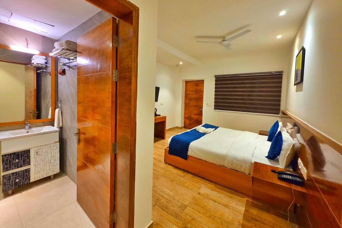 A hotel room with a wooden floor, a bed with a blue and white bedspread, a bedside table with a phone, a ceiling fan, and a bathroom with a wooden door and a sink.